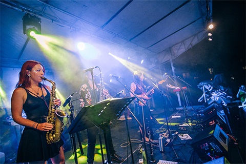 Stage lit up with person playing sax in foreground, and guitarists and drummers behind and to the side